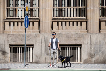 Young blind man with white cane and guide dog standing on pavement in city, waiting. - HPIF17686