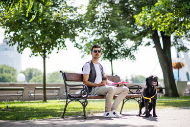 Junger blinder Mann mit weißem Stock und Blindenhund sitzt auf einer Bank in einem Park in der Stadt und ruht sich aus. - HPIF17696