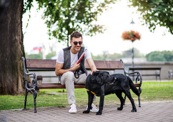 Junger blinder Mann mit weißem Stock und Blindenhund sitzt auf einer Bank in einem Park in der Stadt und ruht sich aus. - HPIF17697