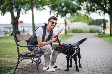 Junger blinder Mann mit weißem Stock und Blindenhund sitzt auf einer Bank in einem Park in der Stadt und ruht sich aus. - HPIF17699