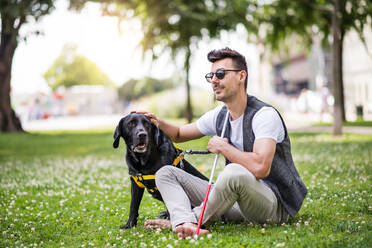 Junger blinder Mann mit weißem Stock und Blindenhund sitzt im Park in der Stadt und ruht sich aus. - HPIF17704