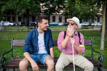 Ein junger Mann und ein blinder Senior mit weißem Stock sitzen auf einer Bank in einem Park in der Stadt und unterhalten sich. - HPIF17719
