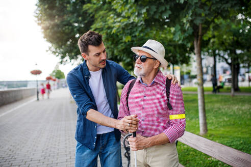 Ein junger Mann und ein blinder Senior mit weißem Stock gehen in der Stadt auf dem Bürgersteig und unterhalten sich. - HPIF17720