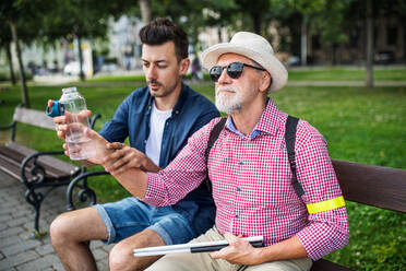 Ein junger Mann und ein blinder Senior mit weißem Stock und Wasserflasche sitzen auf einer Bank in einem Park in der Stadt. - HPIF17722