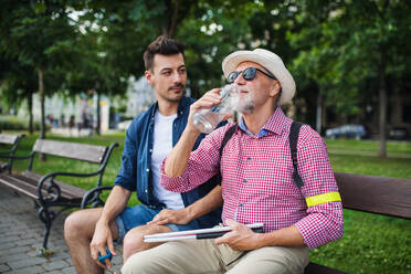 Ein junger Mann und ein blinder Senior mit weißem Stock sitzen auf einer Bank in einem Park in der Stadt und trinken Wasser. - HPIF17723