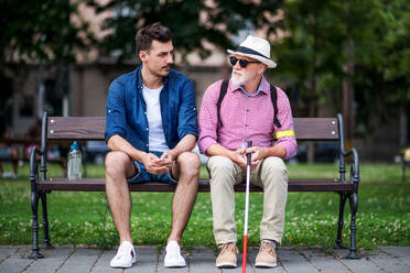 Ein junger Mann und ein blinder Senior mit weißem Stock sitzen auf einer Parkbank in der Stadt. - HPIF17725