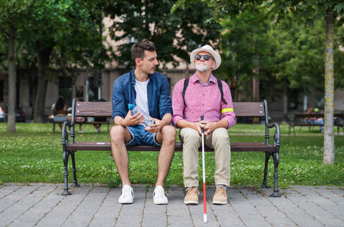 A young man and blind senior with white cane sitting on bench in park in city, talking. - HPIF17726