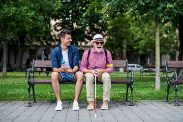 Ein junger Mann und ein blinder Senior mit weißem Stock sitzen auf einer Parkbank in der Stadt. - HPIF17727