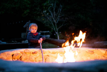 Ein kleiner Junge steht und spielt am offenen Feuer am Abend draußen im Garten im Sommer. - HPIF17826