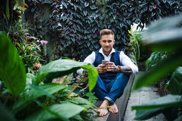 Young man sitting barefoot in botanical garden, using smartphone. Green bussiness concept. - HPIF17883