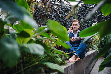 Young man with smartphone sitting in botanical garden, making phone call. Green bussiness concept. - HPIF17884