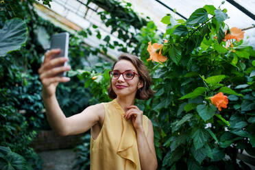 Front view of young woman with smartphone standing in botanical garden, taking selfie. - HPIF17927