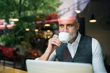 Älterer Mann mit Kaffee am Tisch in einem Café sitzend, Laptop benutzend, Aufnahme durch Glas. - HPIF17958
