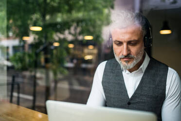 Älterer Mann mit Kopfhörern am Tisch in einem Café, der einen Laptop benutzt, Aufnahme durch Glas. - HPIF17962