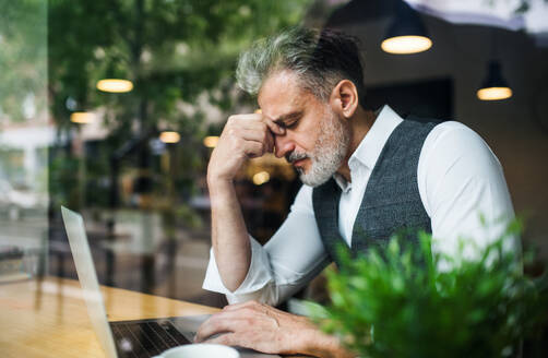 Trauriger und frustrierter reifer Mann, der in einem Café am Tisch sitzt und einen Laptop benutzt, aufgenommen durch Glas. - HPIF17966