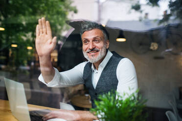 Älterer Mann mit Laptop am Tisch in einem Café, der jemanden begrüßt, Aufnahme durch Glas. - HPIF17967