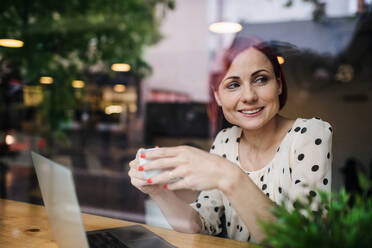 Porträt einer Frau, die mit einem Kaffee am Tisch in einem Café sitzt und einen Laptop benutzt, aufgenommen durch ein Glas. - HPIF17969