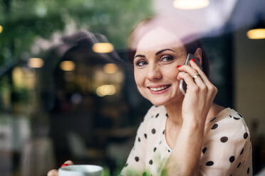 Porträt einer Frau mit Kaffee und Smartphone, die in einem Café am Tisch sitzt und telefoniert, Aufnahme durch Glas. - HPIF17970