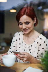 Porträt einer Frau mit Kaffee und Smartphone, die am Tisch in einem Café sitzt und einen Laptop benutzt, aufgenommen durch ein Glas. - HPIF17971