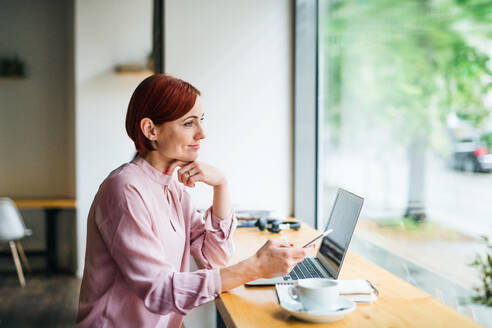 Eine attraktive Frau mit Kaffee, Laptop und Telefon sitzt am Tisch eines Cafés. - HPIF17996