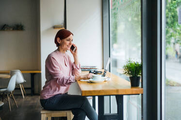 Eine attraktive Frau mit Kaffee und Telefon sitzt am Tisch eines Cafés und telefoniert. - HPIF17998