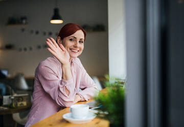 Ein Porträt einer attraktiven Frau, die mit einem Kaffee am Tisch in einem Café sitzt und jemandem zuwinkt. - HPIF18001