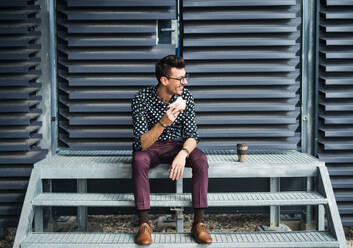 Ein junger Geschäftsmann sitzt mit Kaffee und Sandwich im Freien, isst eine Kleinigkeit und ruht sich aus. - HPIF18038