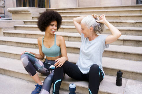 Smiling woman tying hair sitting with daughter on steps - MMPF00765