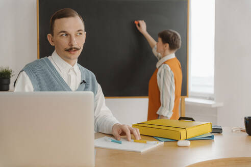 Lächelnder Mann sitzt im Klassenzimmer und schaut auf seinen Laptop, während sein Kollege an die Tafel schreibt - EHAF00024