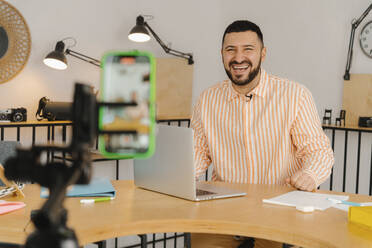 Businessman recording online class with smartphone sitting at table in studio - EHAF00039