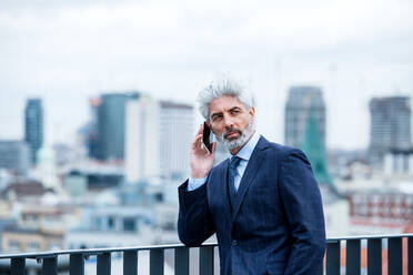 A portrait of mature businessman with smartphone standing on a terrace, working. - HPIF18073