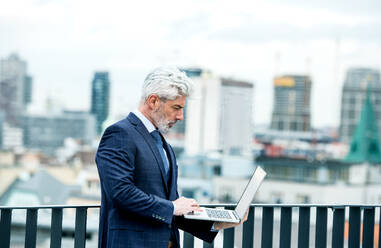 Ein reifer Geschäftsmann mit Laptop steht auf einer Terrasse vor seinem Büro und arbeitet. - HPIF18083