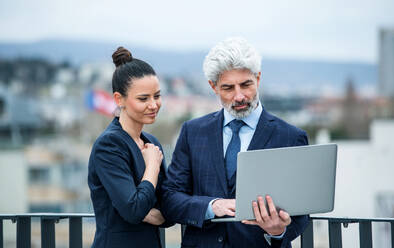 A mature businessman and young businesswoman with laptop standing outdoors on a terrace, working. - HPIF18084