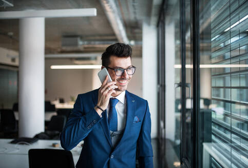 Ein Porträt eines jungen Geschäftsmannes mit Smartphone, der in einem Büro steht und telefoniert. - HPIF18110