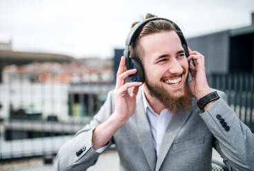 Ein junger Geschäftsmann mit Kopfhörern steht auf einer Terrasse und hört Musik. - HPIF18161