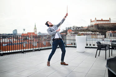 A young businessman with headphones and drumsticks standing on a terrace, having fun. - HPIF18165