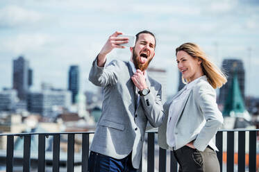 Zwei junge Berufstätige fangen einen Moment der Freude auf einer Terrasse ein, indem sie ein Smartphone benutzen, um ein Selfie vor ihrem Büro zu machen - HPIF18171