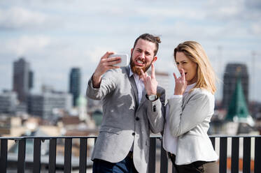 Two cheerful young business people with smartphone standing on a terrace outside office, taking selfie. - HPIF18172