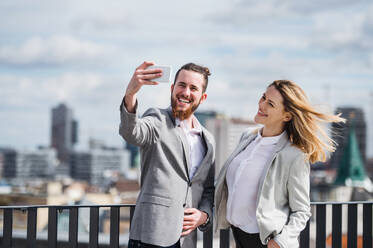 Zwei junge Berufstätige fangen einen Moment der Freude auf einer Terrasse ein, indem sie ein Smartphone benutzen, um ein Selfie vor ihrem Büro zu machen - HPIF18173