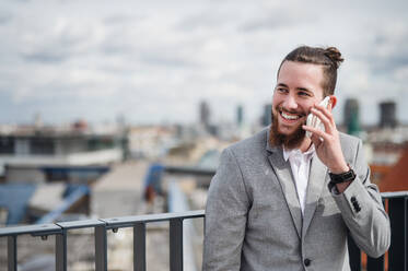 Ein junger Geschäftsmann mit Smartphone steht auf einer Terrasse vor seinem Büro und führt ein Telefongespräch. - HPIF18182