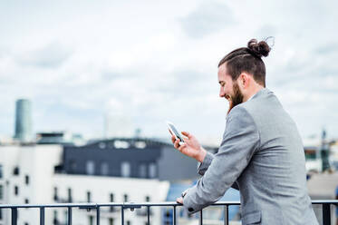 A young businessman with smartphone standing on a terrace outside office, text messaging. Copy space. - HPIF18183