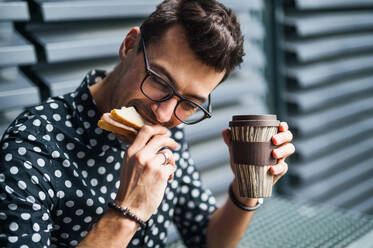 Ein junger Geschäftsmann sitzt im Freien, trinkt Kaffee und isst ein Sandwich. - HPIF18193