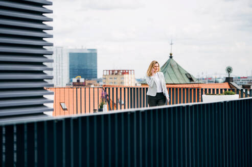Eine junge Geschäftsfrau mit Smartphone steht auf einer Terrasse und führt ein Telefongespräch. - HPIF18196