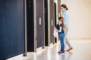 Seitenansicht einer Geschäftsfrau mit kleiner Tochter, die in einem Bürogebäude bei den Aufzügen steht. - HPIF18222