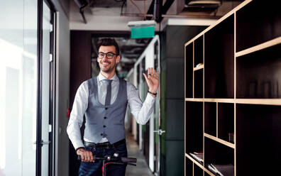 Young businessman with motor scooter in an office building, waving at somebody. - HPIF18262