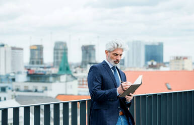 Ein reifer Geschäftsmann mit Tagebuch steht auf einer Terrasse vor seinem Büro und schreibt und plant. - HPIF18286