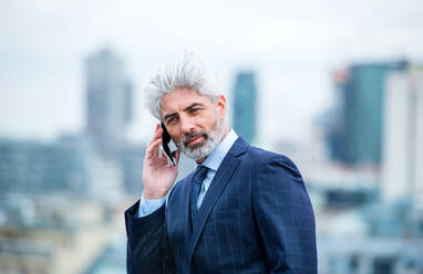 Ein Porträt eines reifen Geschäftsmannes mit Smartphone, der auf einer Terrasse steht und arbeitet. - HPIF18287