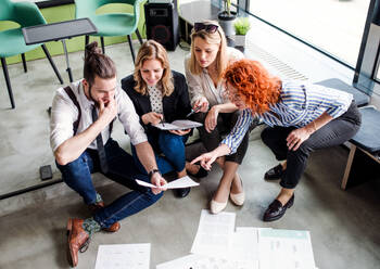 Eine Gruppe junger Geschäftsleute mit einem Terminkalender, die in einem Büro auf dem Boden sitzen und sich unterhalten, Ansicht von oben. - HPIF18343