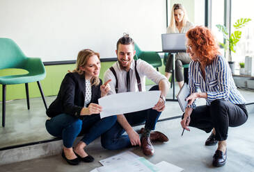 A group of young business people with a diary sitting on the floor in an office, talking. - HPIF18354