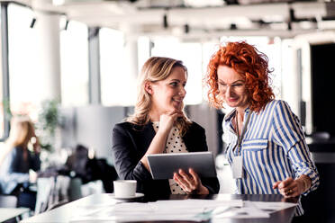 Two cheerful businesswomen with tablet sitting in an office, talking. - HPIF18376